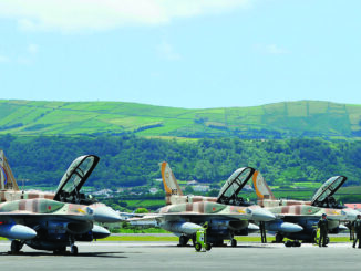 Três caças F-16 da Força Aérea Israelense alinhados em pista de aeroporto com pilotos e técnicos próximos, com colinas verdes ao fundo.