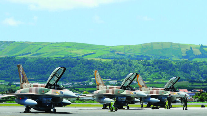 Caças F-16 da Força Aérea Israelense em Base Aérea Três caças F-16 da Força Aérea Israelense alinhados em pista de aeroporto com pilotos e técnicos próximos, com colinas verdes ao fundo.