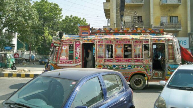 Transporte Tradicional em Karachi Ônibus decorado colorido circulando em rua urbana de Karachi, Paquistão, com carros e edifícios ao fundo.