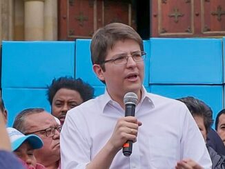 Miguel Uribe Turbay falando em um comício público, segurando microfone, vestindo camisa branca e óculos.