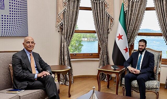 ahmed-al-sharaa-encontro-tom-barrack-maio-2025 Ahmed al-Sharaa em reunião com Tom Barrack, maio de 2025, com a bandeira da Síria ao fundo.