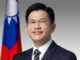 Retrato oficial de Lin Chia-lung, Ministro das Relações Exteriores de Taiwan, vestindo terno escuro e gravata, com a bandeira de Taiwan ao fundo.