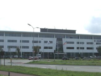 Prédio moderno da sede da ASML em Veldhoven, Holanda, com fachada branca e janelas de vidro, mostrando bandeiras na frente e carros estacionados ao redor.