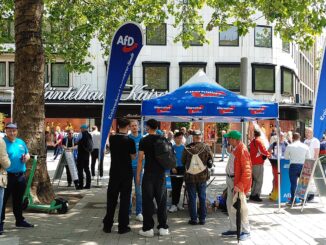 Stand informativo do partido AfD em Hannover com manifestação de oposição ao fundo, ilustrando crescimento da extrema-direita na Alemanha.