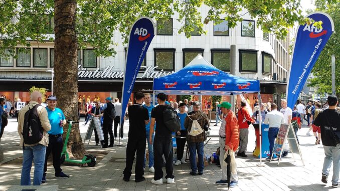 Stand informativo do partido AfD em Hannover com manifestação de oposição ao fundo, ilustrando crescimento da extrema-direita na Alemanha.