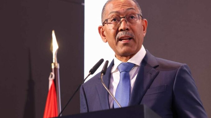 Diamantino Pedro Azevedo em Discurso Oficial Ministro Diamantino Pedro Azevedo de Angola falando em um púlpito durante evento oficial, usando terno azul, gravata azul clara e óculos, com microfones à frente e bandeira de Angola ao fundo.