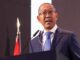 Ministro Diamantino Pedro Azevedo de Angola falando em um púlpito durante evento oficial, usando terno azul, gravata azul clara e óculos, com microfones à frente e bandeira de Angola ao fundo.