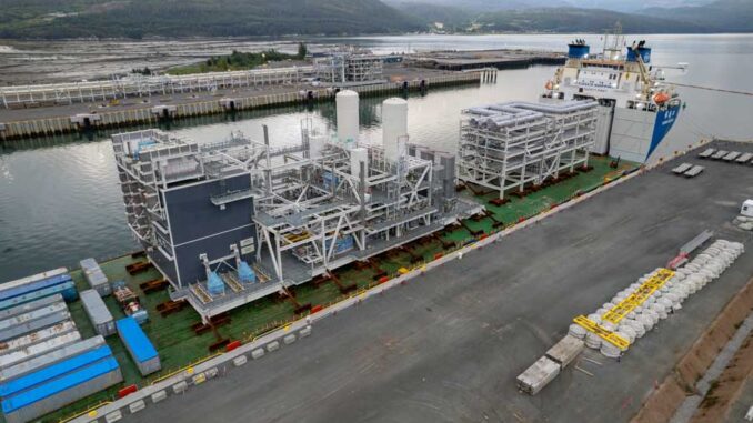 Navio transportando módulos de infraestrutura de GNL em um porto canadense, com montanhas ao fundo e contêineres no cais.