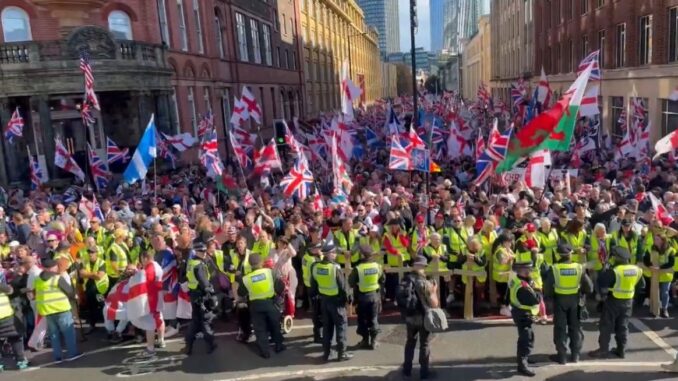 Protesto de direita em Londres 2025 Multidão de manifestantes em Londres segurando bandeiras britânicas, cruzes de São Jorge e bandeiras de países estrangeiros, com policiais em coletes amarelos formando barreiras à frente.