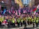 Multidão de manifestantes em Londres segurando bandeiras britânicas, cruzes de São Jorge e bandeiras de países estrangeiros, com policiais em coletes amarelos formando barreiras à frente.