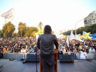 Javier Milei de costas falando para uma multidão em comício político ao ar livre, com bandeiras e público atento em Buenos Aires.