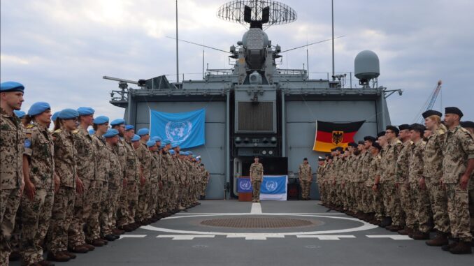Soldados da Bundeswehr em uniforme de camuflagem alinhados no convés de um navio, com bandeiras da Alemanha e da ONU ao fundo, participando de missão da Força-Tarefa Marítima UNIFIL no Líbano.