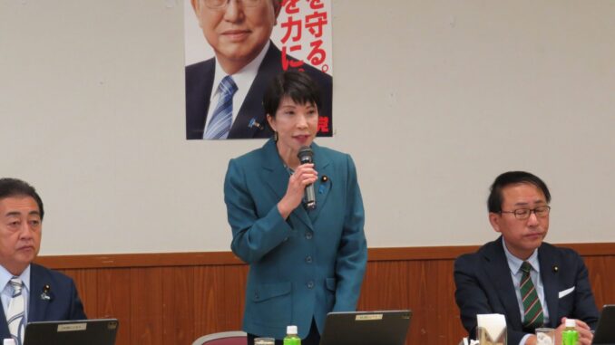 Sanae Takaichi falando em reunião do Grupo de Estudos de Segurança Pública, Terrorismo e Combate ao Crime Cibernético do PLD, com membros do partido e laptops à mesa.