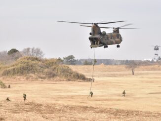 Helicóptero militar japonês transporta soldados por corda durante exercício no Acampamento Narashino, com tropas correndo no campo e outros helicópteros ao fundo.