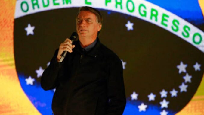 Jair Bolsonaro falando ao microfone com a bandeira do Brasil ao fundo.