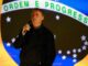 Jair Bolsonaro falando ao microfone com a bandeira do Brasil ao fundo.