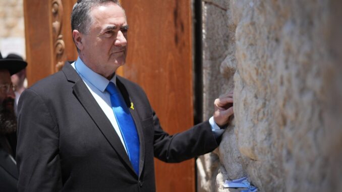 Israel Katz no Muro das Lamentações O ministro da Defesa de Israel, Israel Katz, vestido com terno preto e gravata azul, encosta a mão no Muro das Lamentações em Jerusalém, enquanto mensagens de papel estão inseridas entre as pedras.