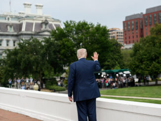 Presidente Donald Trump de costas, acenando para um grupo de jornalistas em frente a prédios governamentais em Washington, EUA.