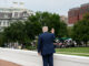 Presidente Donald Trump de costas, acenando para um grupo de jornalistas em frente a prédios governamentais em Washington, EUA.