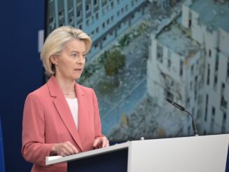 Ursula von der Leyen, presidente da Comissão Europeia, falando em frente a um microfone, com a bandeira da União Europeia ao fundo e imagens de destruição de prédios em tela atrás dela.