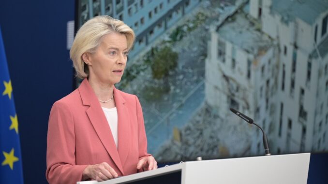 Von der Leyen anuncia planos de envio de tropas à Ucrânia Ursula von der Leyen, presidente da Comissão Europeia, falando em frente a um microfone, com a bandeira da União Europeia ao fundo e imagens de destruição de prédios em tela atrás dela.
