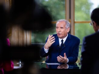 Primeiro-ministro francês François Bayrou em entrevista, usando terno azul e gravata, gesticulando com as mãos enquanto fala, com pessoas parcialmente visíveis ao fundo.