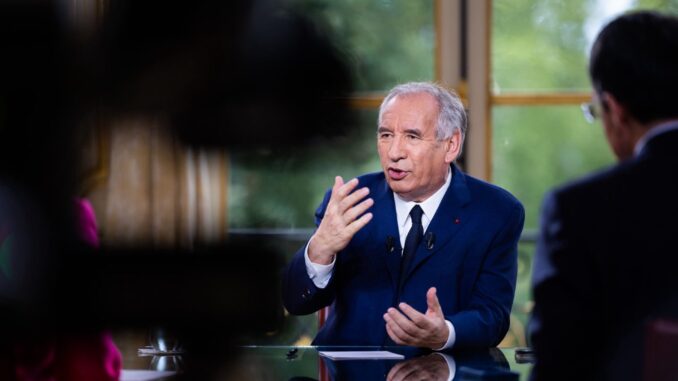 Crise Política na França 2025 Primeiro-ministro francês François Bayrou em entrevista, usando terno azul e gravata, gesticulando com as mãos enquanto fala, com pessoas parcialmente visíveis ao fundo.