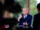 Primeiro-ministro francês François Bayrou em entrevista, usando terno azul e gravata, gesticulando com as mãos enquanto fala, com pessoas parcialmente visíveis ao fundo.