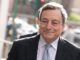 Mario Draghi sorrindo, vestindo terno escuro e gravata, em evento público ao ar livre.