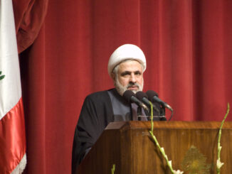 Naim Qassem, líder do Hezbollah, discursando em um púlpito com a bandeira do Líbano ao fundo.