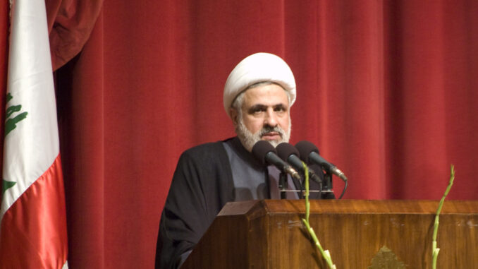 Naim Qassem, líder do Hezbollah, discursando em um púlpito com a bandeira do Líbano ao fundo.