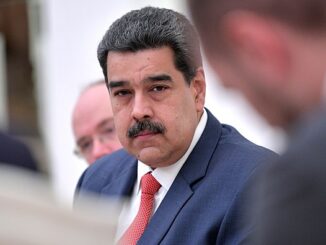 Nicolás Maduro em traje formal olhando para frente durante uma reunião, com outras pessoas desfocadas ao fundo.